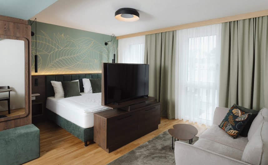 Einladendes Schlafzimmer mit dekorativer Wandgestaltung, Fernseher, Sofa und gro&szlig;en, hellen Fenstern.